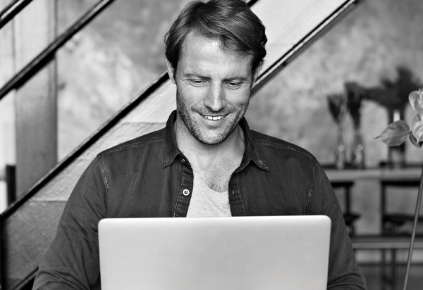 male sitting holding computer smiling at screen. Photo is in black and white.